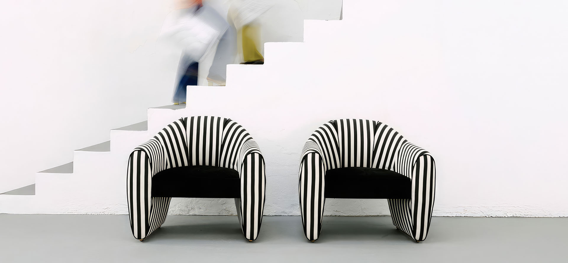 Viginti XX Armchair Two Viginti XX armchairs upholstered in striped black and white fabric, placed near a staircase with two out-of-focus figures ascending.