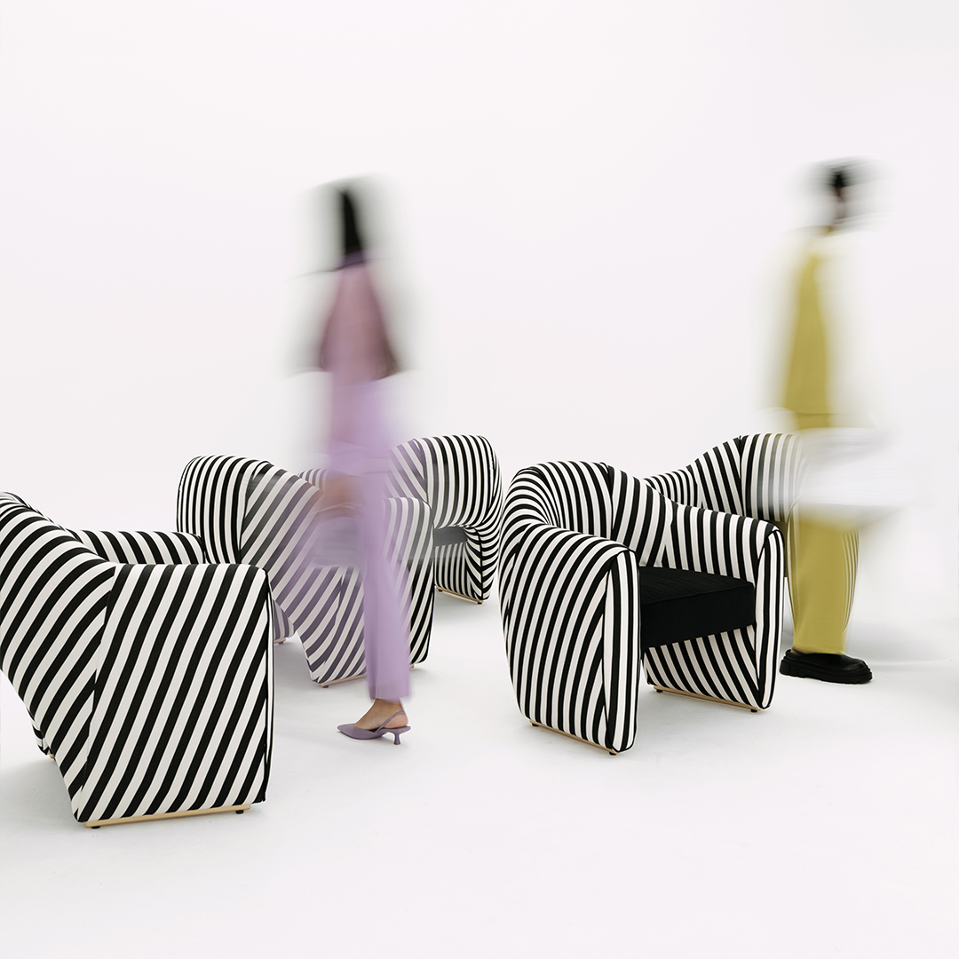 Viginti XX Armchair Five Viginiti XX armchairs in a studio setting, upholstered in black and white striped fabric, with two people walking by in the background, out of focus, talking about the brand.