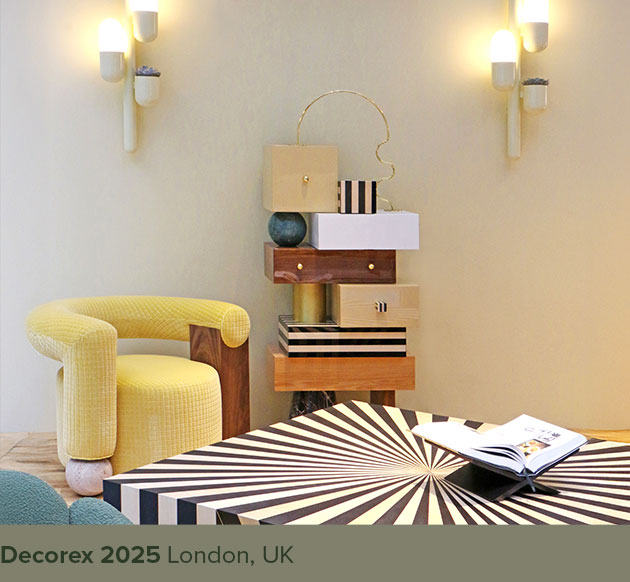 Galatea armchair upholstered in yellow checkered fabric beside the Sketch chest of drawers, with an Op-Art center table in the foreground. Two illuminated Cactus wall lamps cast a warm yellow glow across the scene, showcased at Decorex 2025.