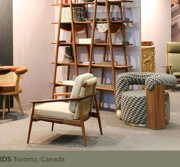 IDS Toronto A view of a furniture exhibition showcasing the "Adam armchair", "Galatea armchair", "Blake shelf", and "George dining chair" on a gray carpet. In the foreground sits the Adam armchair, featuring a dark wood frame, light-colored upholstery, and a woven cane backrest. To the right, the rounded Galatea armchair is upholstered in a black-and-white geometric pattern, accented by a solid wood block on its side. In the background stands the tall, open-frame wooden Blake shelf with shelves displaying various items, and to the far left, the George dining chair is partially visible. At the bottom of the image, a gray-green banner displays the text: "IDS Toronto, Canada".