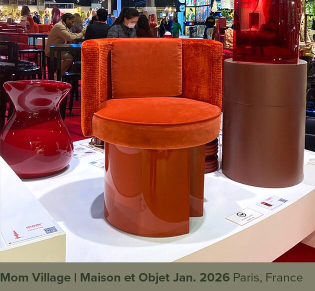 MOM Village A modern design chair named "Myth" in an orange-terracotta tone, featuring a round velvet seat and a curved backrest, supported by a glossy cylindrical base. To the left of the "Myth" chair is a glossy red, wavy ceramic vase, and to the right is a brown column-like pedestal. The background shows a bustling design fair with people and other exhibits. At the bottom of the image, a gray-green banner displays the text: "Mom Village | Maison et Objet Jan. 2026 Paris, France".
